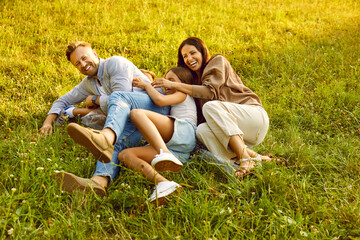 Fototapeta premium Happy young family of four lying on green grass and having fun in summer park. Smiling parents hugging and playing with kids boy and girl in nature enjoying time together. Family leisure concept.
