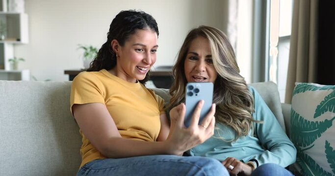 Mature woman with young adult daughter sitting on sofa looking at smart phone screen laugh at funny videos, watch on-line, spend weekend on internet. Leisure of family at home using modern technology