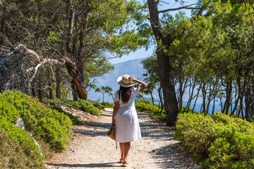 Rear view of woman wearing white sun dress standing on gravel path under pine trees by sea shore - obrazy, fototapety, plakaty
