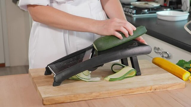 Chef's hands grate green zucchini with slices on a mandalin grater, wooden table on a background of vegetables, home cooking, vegetarian food, vegetables rich in vitamins, diet. Close-up, side view