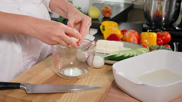 In the kitchen, the filling for the zucchini and feta pie is being prepared. The chef breaks eggs into a glass measuring cup on a wooden board, with vegetables in the background. Close-up, side view.