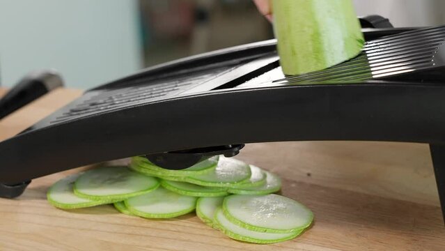 The chef on a wooden table grates green zucchini and zucchini into circles on a mandalin grater. Close-up, front view, macro