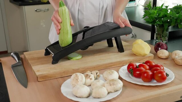 The chef puts a magdalene grater on a wooden table and grates zucchini, zucchini into circles, a wooden table against a background of vegetables. Close-up, front view