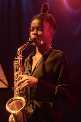 Vertical portrait of young Black woman playing saxophone performing on stage with jazz band