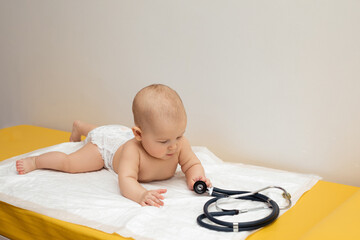 A little baby is lying on the couch in a diaper and playing with a stethoscope. 