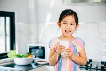Asian preschooler enjoys milk in kitchen smiling happily. Portrait of cute daughter holding drink radiating joy. Happy little girl sips calcium-rich liquid feeling cheerful at home give me.