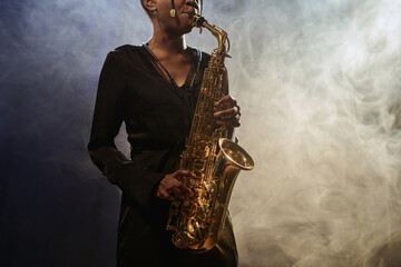 Cropped shot of elegant Black young woman playing saxophone in jazz music club with smoke effects...