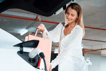 Young woman travel with EV electric car to shopping center parking lot charging in downtown city...