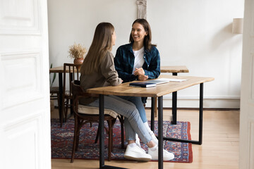 Happy young female colleagues discussing work projects in stylish co-working apartment, sitting at table in loft interior, talking, smiling, laughing. Wide shot through doorway view
