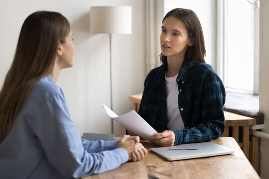 Serious young project manager woman in casual informal clothes talking to female business colleague at work table, using paper documents, holding resume, interviewing job candidate,