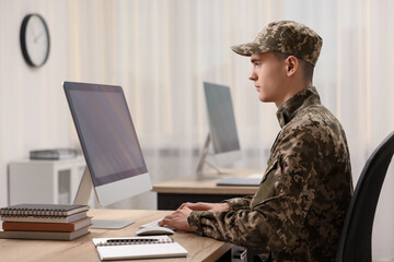 Military service. Young soldier working with computer at wooden table in office - obrazy, fototapety, plakaty