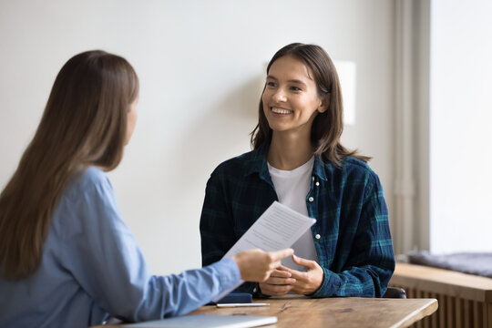 Happy young job candidate woman in informal clothes talking to employee on interview, talking to boss with toothy smile, meeting with colleague at work table, discussing paper resume