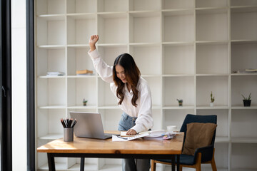 Cheerful young businesswoman raising her arm in a victory gesture at her office desk.
