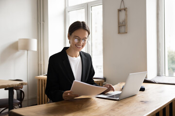 Positive confident financial specialist, accountant, lawyer woman reviewing papers at workplace, reading legal documents, sales reports, using laptop, smiling, laughing, getting good news