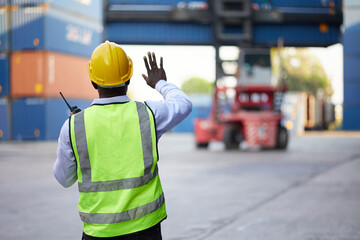 back view African worker or engineer using walkie talkie and showing gesture to crane car in containers warehouse storage