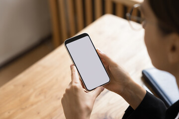 Blank screen of smartphone in young business woman hands. Female employee, manager using mobile phone, holding gadget over workplace office, looking at blank white touch screen