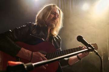 Low angle portrait of long haired mature musician playing acoustic guitar on stage and singing to...