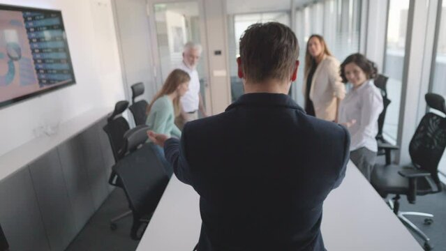 Company CEO standing and letting employees to sit down in boardroom, back view slow motion. Executive starting business meeting with corporate workers in conference room, showing leadership concept
