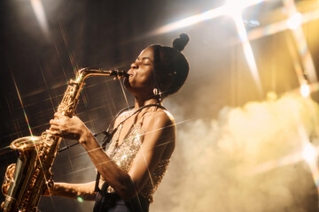 Portrait of talented Black young woman playing saxophone performing jazz music at concert in golden lights copy space - obrazy, fototapety, plakaty