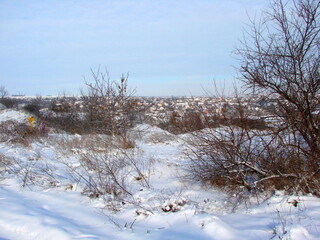 A view from the top of a snow-covered hill to the private houses of the settlement under a clear blue-white sky on the horizon.
