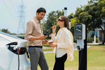 Young couple with coffee pay for electricity while recharge EV car battery at charging station...