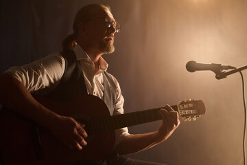 Side view portrait of classic mature guitarist playing acoustic guitar during stage performance in soft spotlight, copy space