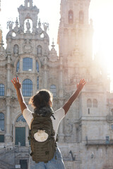 A woman, seen from behind, wearing a backpack adorned with a scallop shell, raising her arms in a gesture of victory and enjoyment, with the Santiago de Compostela Cathedral in the background