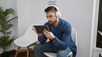 Relaxed young hispanic man enjoying tech-life in waiting room, sitting with touchpad, immersed in music through headphones