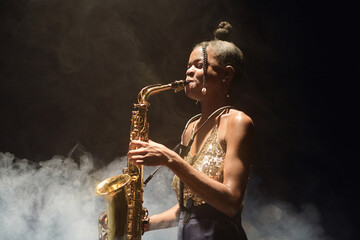 Side view portrait of young Black woman playing saxophone and performing jazz music with smoke in background, copy space - obrazy, fototapety, plakaty