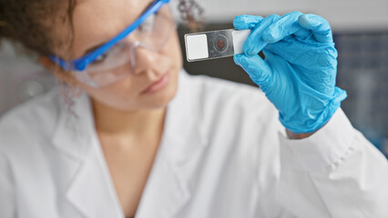 A focused young hispanic woman in a lab coat examines a slide while working indoors at a laboratory.