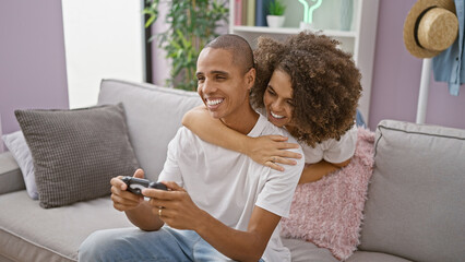 Beautiful couple hugging, full of love, playing video game together at home. confident in their relationship, they enjoy their happy lifestyle, sitting on the sofa, full of smiles. - obrazy, fototapety, plakaty
