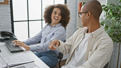 Two team partners, a man and a woman, enjoy working together at the office, smiling confidently while using a computer in indoor professional surroundings