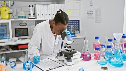 In the heart of the lab, attractive african american woman scientist seriously taking notes, working with microscope in bustling medical research center - obrazy, fototapety, plakaty