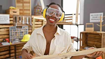 Beautiful african american woman carpenter, bedecked in security glasses and headphones, masterfully handling wood plank in bustling carpentry workshop
