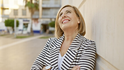 Mature caucasian woman standing outdoors in a city, smiling while leaning against a wall dressed in striped attire.