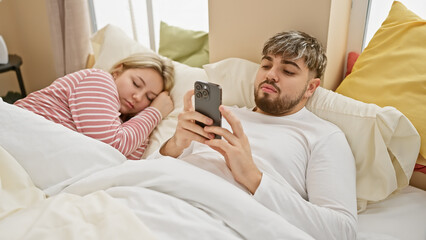 A man using a smartphone while a woman sleeps beside him in a cozy bedroom setting, depicting a modern couple's lifestyle.