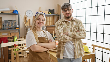 Confident man and woman team standing together in a sunny carpentry workshop full of tools and wood.