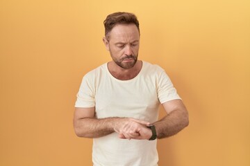 Middle age man with beard standing over yellow background checking the time on wrist watch, relaxed and confident