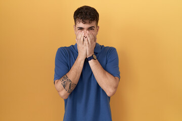 Young hispanic man standing over yellow background laughing and embarrassed giggle covering mouth...