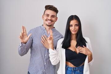 Young hispanic couple standing over white background smiling swearing with hand on chest and...