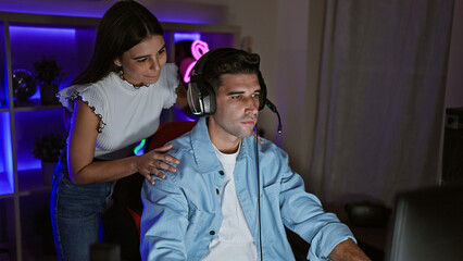 A woman leans affectionately over a man gaming at night in a cozy, darkened room lit by colorful led lights.