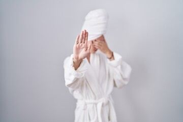 Blonde caucasian woman wearing bathrobe covering eyes with hands and doing stop gesture with sad...