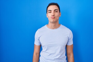 Fototapeta premium Young hispanic man standing over blue background looking at the camera blowing a kiss on air being lovely and sexy. love expression.