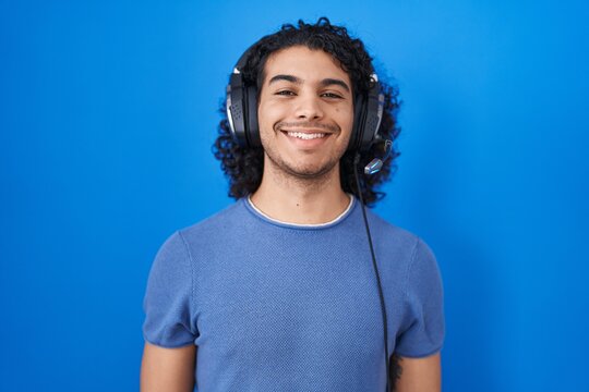 Hispanic man with curly hair listening to music using headphones with a happy and cool smile on face. lucky person.