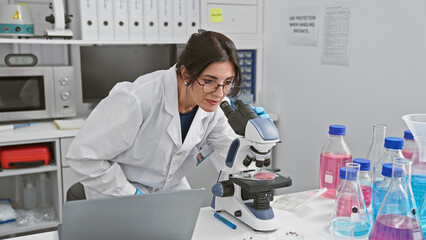 Mature hispanic woman scientist analyzing samples with microscope in laboratory indoor setting.