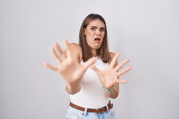 Hispanic young woman standing over white background afraid and terrified with fear expression stop gesture with hands, shouting in shock. panic concept.