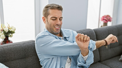 A handsome hispanic man stretches while sitting on a grey sofa in a bright, cozy living room.