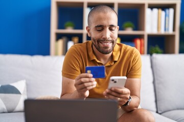 Young latin man using smartphone and credit card sitting on sofa at home - obrazy, fototapety, plakaty