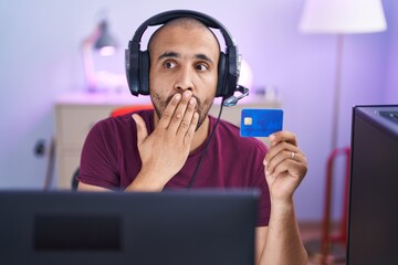 Hispanic man with beard doing online shopping with computer and credit card covering mouth with...