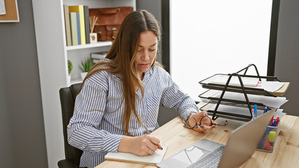 Focused woman writing notes in a modern office setting, with laptop and organized documents.